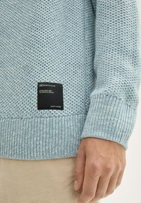 Hellblaue, strukturierte Strickjacke mit gerippten Bündchen und Saum. Mit einem schwarzen Markenlabel, das an der linken Seite nahe dem Saum eingestickt ist.