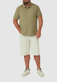 Mann trägt ein olivgrünes Poloshirt, beige knielange Shorts und weiße Sneakers, steht mit einer Hand in der Tasche.