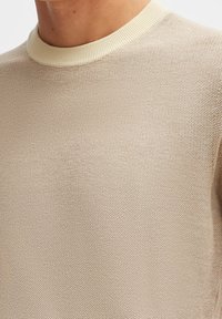 Pull en maille beige clair avec un motif texturé, présentant un col côtelé crème. Le modèle est à col rond et manches courtes.