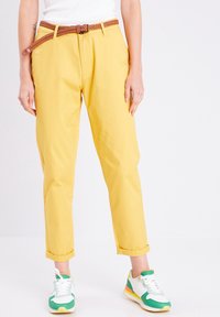 Pantalons en coton jaunes avec une taille ceinturée, accompagnés d'une ceinture en cuir tressé marron. Manches retroussées et poches, associés à des baskets vertes et blanches.