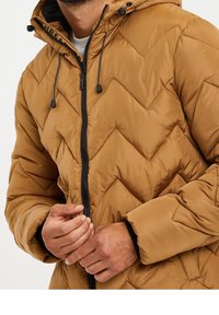 Man draagt een bruine gewatteerde jas met een zwarte rits en verstelbare capuchon, terwijl hij de ritssluiting over een lichtgrijs shirt omhoog trekt.