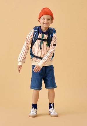 Child wearing a cream and red striped long-sleeve shirt, denim shorts, white sneakers, and an orange beanie, with a blue backpack.