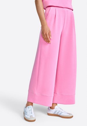 Person, die weite pinke Hosen und weiße Sneakers mit grauen Streifen auf einem einfarbigen hellen Hintergrund trägt.