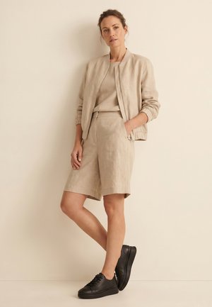 Žena se opírá o béžovou zeď, má na sobě béžovou bomber bundu, odpovídající kraťasy a černé tenisky, ruce v kapse.