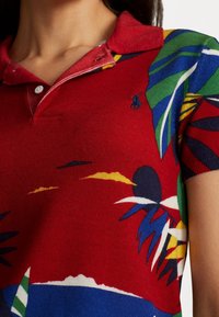 Nära beskjud av en röd polo skjorta med flerfärgade tropiska blad och abstrakta mönster, med en liten marinblå broderad logotyp på bröstet.