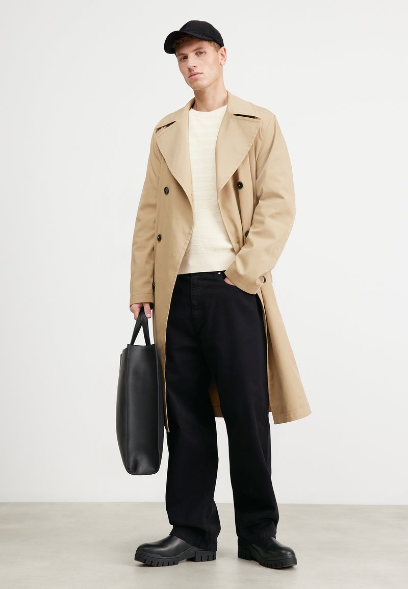 Beżowy płaszcz trenczowy z dużymi klapami i podwójnymi guzikami, kremowy sweter, czarne spodnie, masywne czarne botki i czarna torba na ramię.
