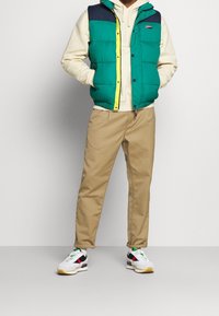 Chaleco verde acolchado con sección superior azul marino, detalle de cremallera amarilla, pantalones beige y zapatillas blancas con acentos negros y rosas.