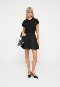 Black short-sleeve top with embroidered designs, paired with a black skirt featuring a gathered waist and polka dot pattern; holding a black handbag.