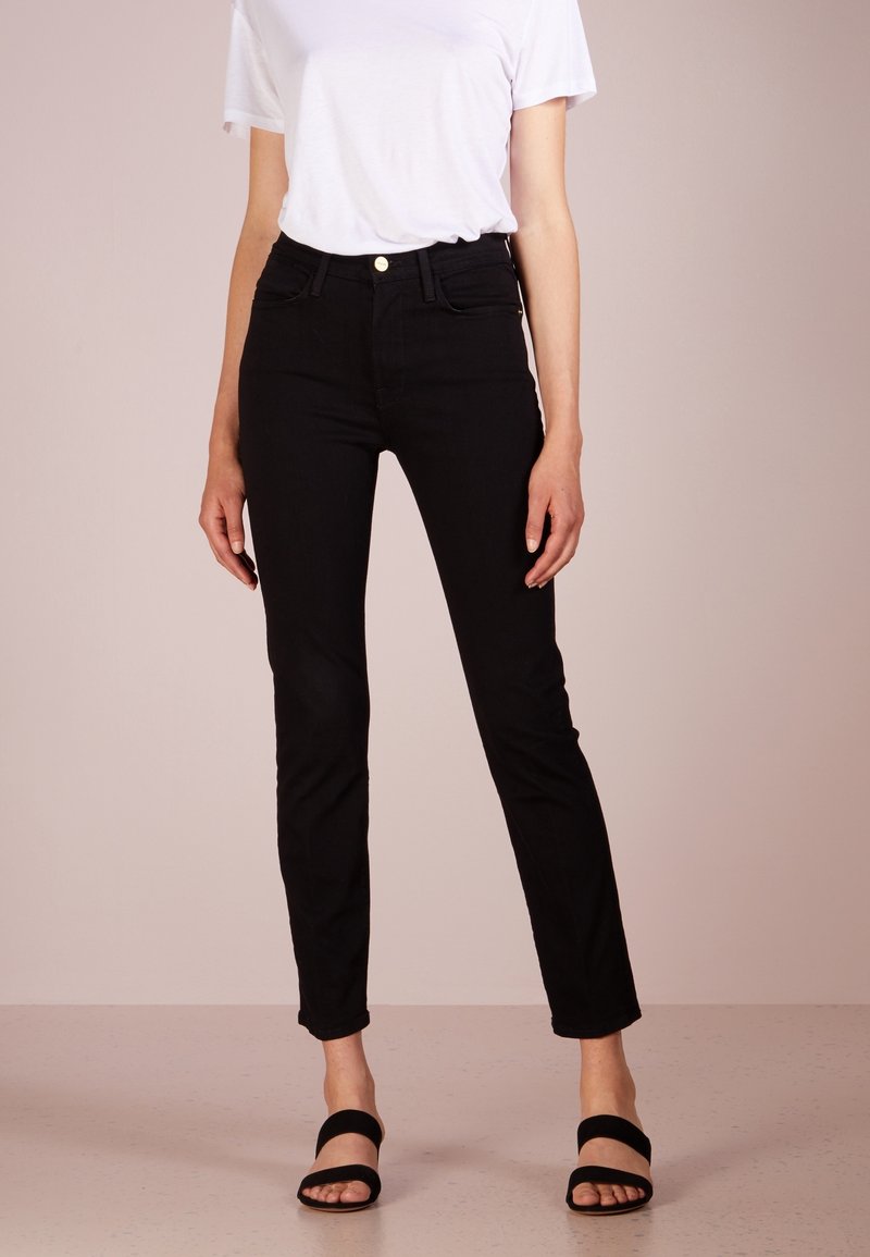 Femme portant un jean noir ajusté, un t-shirt blanc à manches courtes et des sandales à talons noires ouvertes à l'avant, debout devant un fond neutre.