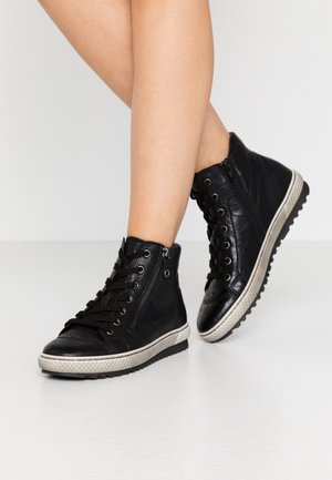 Osoba nosząca czarne wysokie skórzane sneakersy z sznurowadłami i bocznymi suwakami, stojąca na jasnej podłodze.