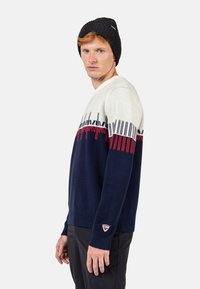 Marineblau und cremefarbener Strickpullover mit roten Akzenten, versehen mit geometrischen Mustern und gerippten Bündchen. Kombiniert mit einer schwarzen Strickmütze.