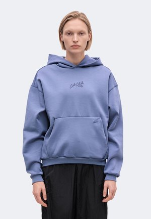 Un mannequin se tient face à l’objectif, vêtu d’un sweat-shirt bleu à capuche avec une poche sur le devant et d’un pantalon noir, sur un fond clair uni.