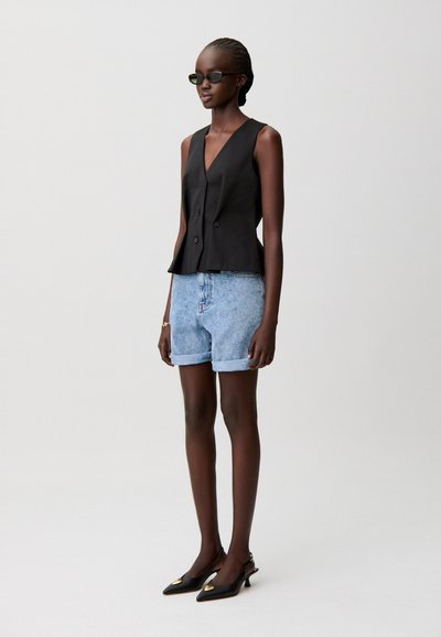 Gilet nero senza maniche con orlo a volant, abbinato a pantaloncini di denim azzurri. Scarpe col tacco slingback nere con dettagli dorati completano il look.