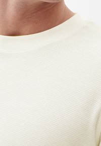 Maglietta leggera e strutturata a maniche lunghe di un color crema chiaro con scollo rotondo e un sottile motivo a coste. Design semplice e casual.
