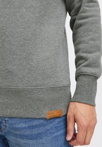 Sudadera gris con tejido texturizado, puños y dobladillo acanalados. Presenta una etiqueta de marca de cuero en la costura inferior. Combinada con jeans azules.