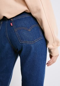 Jeans in denim blu scuro con vestibilità dritta, caratterizzati da cuciture dorate e due tasche posteriori con un design curvo distintivo. Etichetta del marchio rossa visibile.