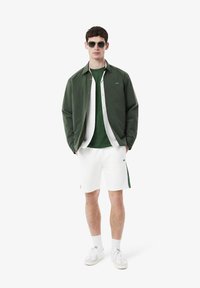 Giacca leggera verde sopra un top verde, pantaloni corti bianchi con striscia verde ai lati e sneakers bianche. Gli occhiali da sole completano l'outfit.