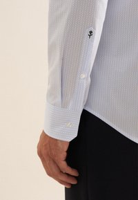 Camicia button-up azzurro chiaro con un piccolo motivo circolare, dotata di polsini bianchi e un logo ricamato nero sulla manica.