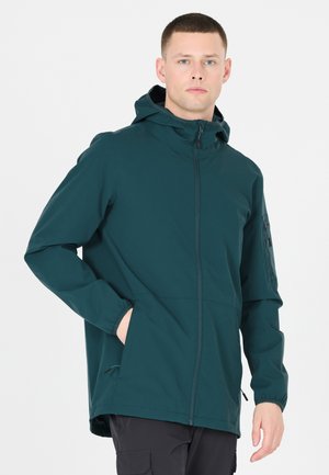 Joven con chaqueta oscura con capucha y bolsillos delanteros y pantalones cargo negros, mirando hacia adelante sobre un fondo liso.