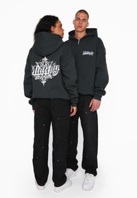 Felpa con zip di colore grigio scuro con una grafica bianca sulla schiena e sul petto, abbinata a jeans neri e sneakers bianche. Vestibilità comoda.