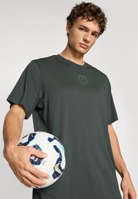 Mørkegrøn bomulds T-shirt med rund halsudskæring og korte ærmer, der har et subtilt cirkulært logo på brystet. En hvid fodbold holdes i hånden.