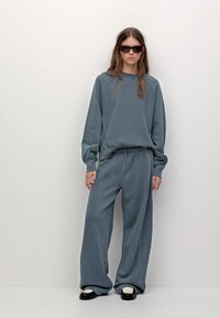 Niebieski, oversize'owy sweatshirt z rękawami raglanowymi, w połączeniu z dopasowanymi spodniami o szerokich nogawkach. Posiada swobodną, miękką fakturę. Podkreślony ciemnymi okularami przeciwsłonecznymi.