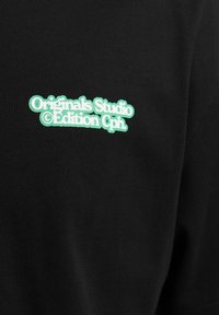 Camiseta de tela negra con texto bordado en verde: "Originals Studio © Edición Cph." Textura suave, diseño de ajuste ceñido.