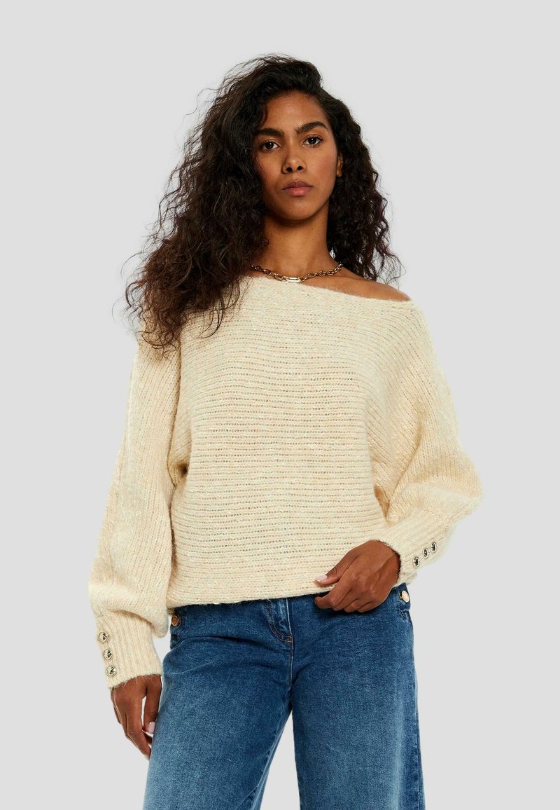 Maglione lavorato a maglia color avorio con una vestibilità over, scollo a barca, spalle scese e dettagli di bottoni sulle maniche. Abbinato a jeans blu.