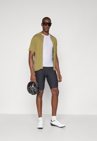 Radfahroutfit mit einem olivgrünen kurzärmligen Trikot, weißem Mesh-Unterhemd, schwarzen Shorts und einem schwarzen Helm. Weiße Radschuhe runden das Outfit ab.