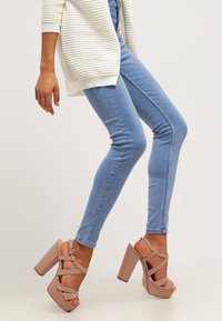 Sandales à plateforme à talons hauts beiges avec de larges brides croisées et des talons épais, associées à un jean skinny bleu clair et un cardigan blanc texturé.