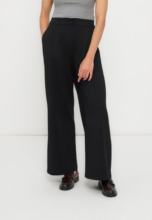 Kvinde iført højtaljede, vide sorte bukser, grå ærmeløs ribbet top og mørkebrune læderloafer, stående med den ene hånd i lommen.