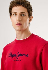 Joven con cabello rizado que lleva una sudadera roja con "Pepe Jeans London" bordado en azul marino en la parte frontal.