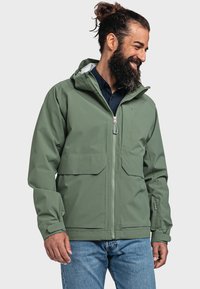 Giacca impermeabile verde con zip frontale, due grandi tasche anteriori, polsini regolabili e cappuccio. Indossata sopra una maglietta scura e jeans.