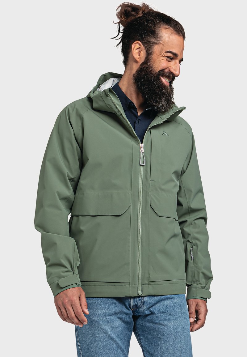Giacca impermeabile verde con zip frontale, due grandi tasche anteriori, polsini regolabili e cappuccio. Indossata sopra una maglietta scura e jeans.