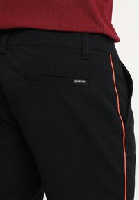 Černé kalhoty s hladkou texturou, s kontrastním oranžovým bočním pruhem a malým štítkem "YOURTURN" na zadní kapse.