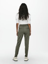 Γυναίκα με μακριά κυματιστά μαλλιά, που φοράει λευκό τοπ με φουσκωτά μανίκια, χακί παντελόνι με ζώνη και λευκά αθλητικά, γυρισμένη πλάτη σε απλό φόντο.