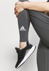 Černá sportovní obuv s bílou podrážkou a texturovaným svrškem, doplněná šedými kompresními legínami s bílým logem a pruhovanými akcenty.
