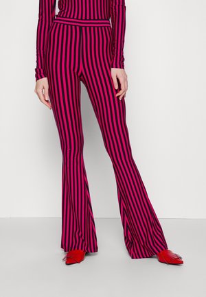 Persoon die roze en zwart verticaal gestreepte uitlopende broek en bijpassende top draagt, gecombineerd met glanzende rode puntige schoenen met franjes.