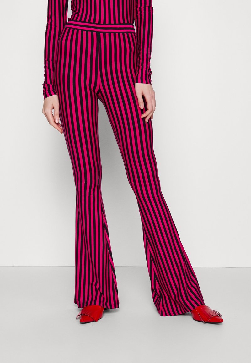 Person trägt rosa-schwarz vertikal gestreifte Schlaghosen und passendes Oberteil, kombiniert mit glänzenden roten spitzen Schuhen mit Quasten.