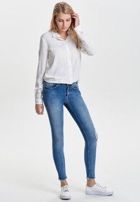 Camisa blanca de botones, jeans ajustados azules con detalles desgastados y zapatillas blancas. Tejido suave, diseño ajustado y variaciones sutiles en el lavado.