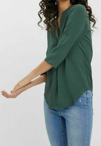 Blouse vert foncé à manches trois-quarts, encolure en V, tissu texturé et coupe décontractée. L'ourlet est légèrement plus long à l'arrière.