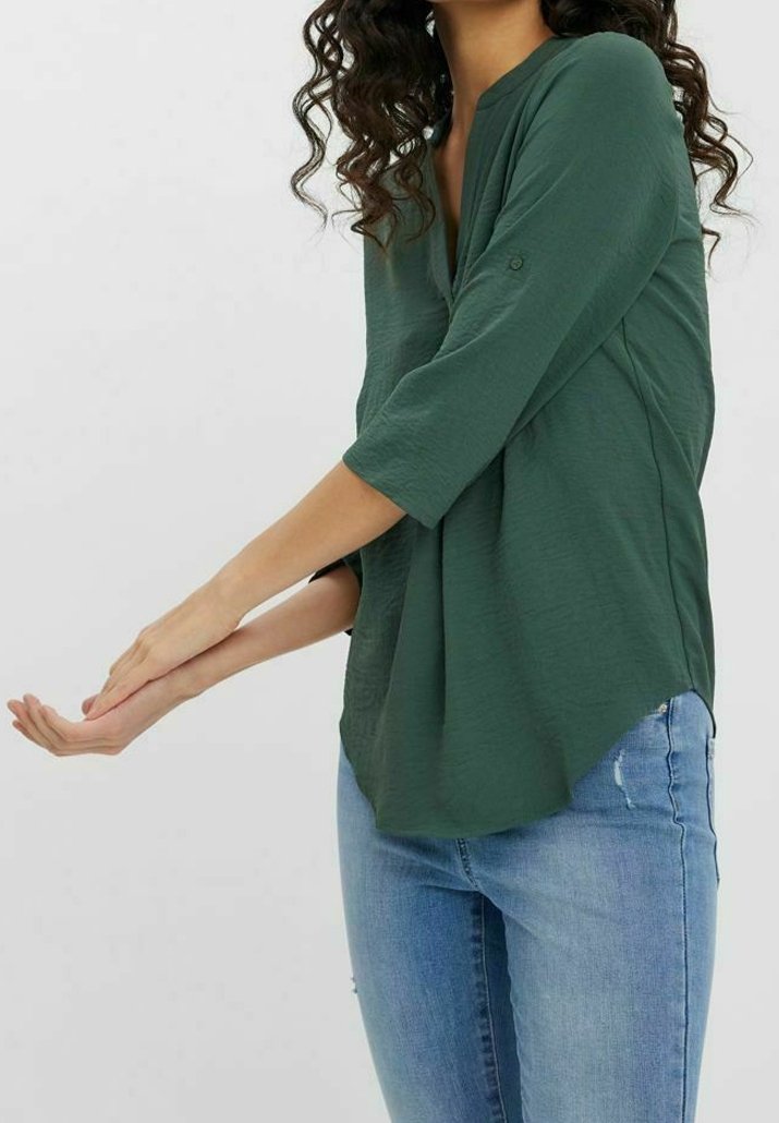 Blouse vert foncé à manches trois-quarts, encolure en V, tissu texturé et coupe décontractée. L'ourlet est légèrement plus long à l'arrière.