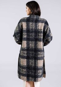 Manteau long à carreaux en laine noire, grise et beige. Doté d'un col montant, de manches oversize et d'une fente sur les côtés pour faciliter les mouvements.