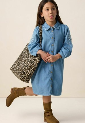 Jeune fille en robe en jean bleu et bottes marron tenant un sac fourre-tout à motifs léopard, tirant la langue de manière ludique avec une jambe levée.