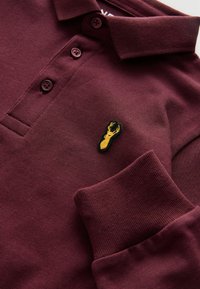 Bordo polo krekls ar teksturētu audumu, īsu apkakli, trim pogām un dzeltenu izšūtu omāru akcentu uz krūtīm.