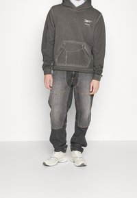 Felpa grigia con cappuccio e tasca frontale caratterizzata da un dettaglio a catena, abbinata a pantaloni in denim scuro strappati e sneakers grigie chiare.