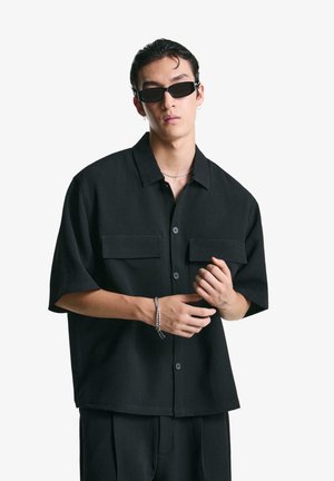 Black short-sleeve shirt with two front pockets, button closure, a relaxed fit, and smooth texture. Worn with black rectangular sunglasses.