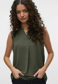 Haut sans manches vert avec un décolleté en V, texture douce et détails de plis subtils, associé à un jean foncé, les mains dans les poches.