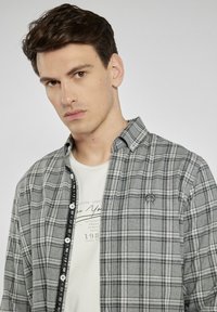 Szara koszula w kratę z zapięciem na guziki, z czarnym akcentem z przodu i małym logo na piersi. Nosi się ją na białym t-shircie.