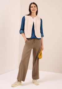 Femme portant une chemise en denim bleu, un gilet en polaire crème, un pantalon large marron, des baskets beiges, tenant un bonnet en tricot moutarde plié, se tenant contre un fond beige.